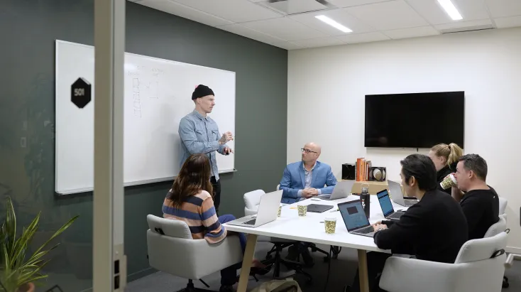 Office meeting with six people, one presenting by a whiteboard.