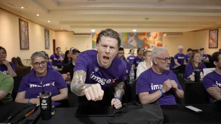Man enthusiastically cheering, seated among others in a conference room.