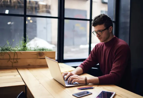 Man in glasses working on a laptop at a wooden desk by a window.