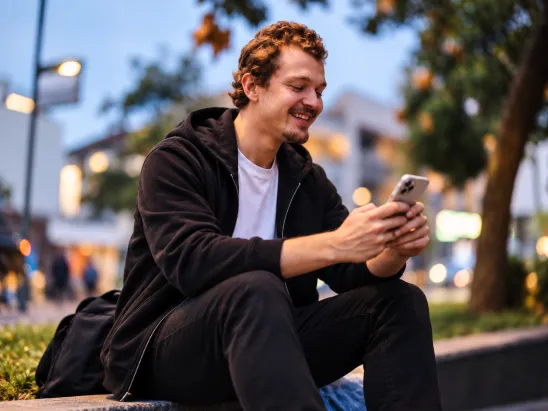 A smiling man sitting outdoors in an urban setting at dusk, using his smartphone, representing mobile-first digital communication and engagement.