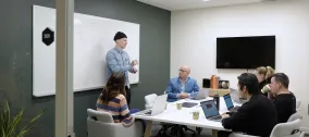 Office meeting with six people, one presenting by a whiteboard.