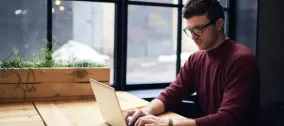 Man in glasses working on a laptop at a wooden desk by a window.