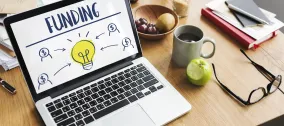 Laptop on a table displaying "Funding" with a light bulb graphic.