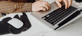 Person working on a laptop at a desk with a mouse and cup of coffee.