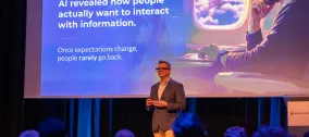 Gleen presenting on stage to a seated audience, with a slide explaining how AI is changing the way people interact with information.