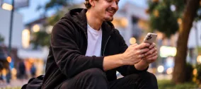 A smiling man sitting outdoors in an urban setting at dusk, using his smartphone, representing mobile-first digital communication and engagement.