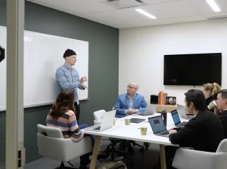 Office meeting with six people, one presenting by a whiteboard.