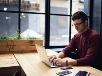 Man in glasses working on a laptop at a wooden desk by a window.