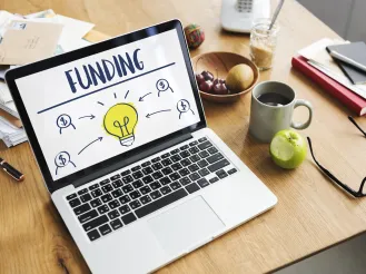 Laptop on a table displaying "Funding" with a light bulb graphic.