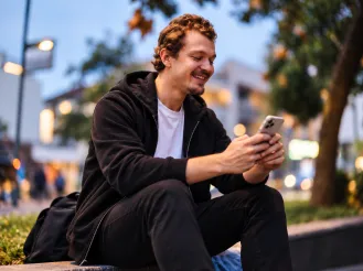 A smiling man sitting outdoors in an urban setting at dusk, using his smartphone, representing mobile-first digital communication and engagement.