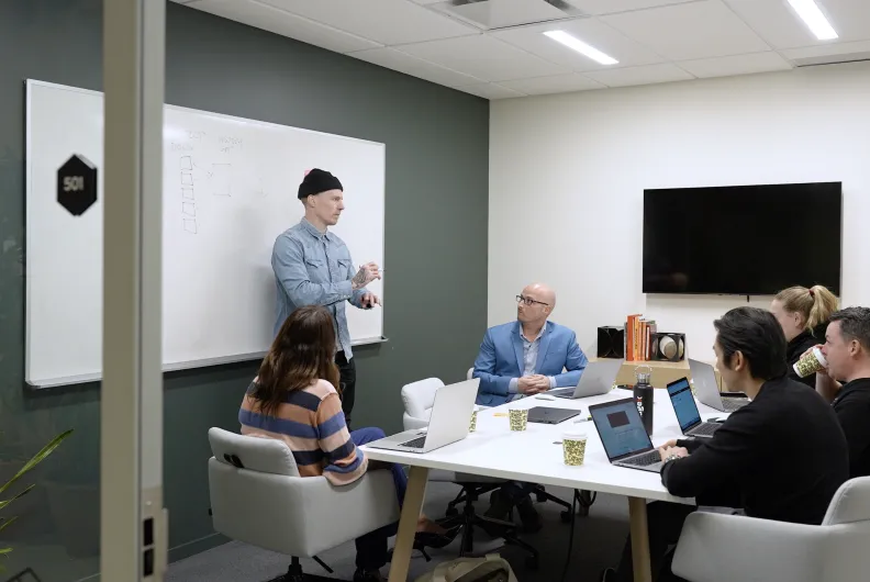 Office meeting with six people, one presenting by a whiteboard.