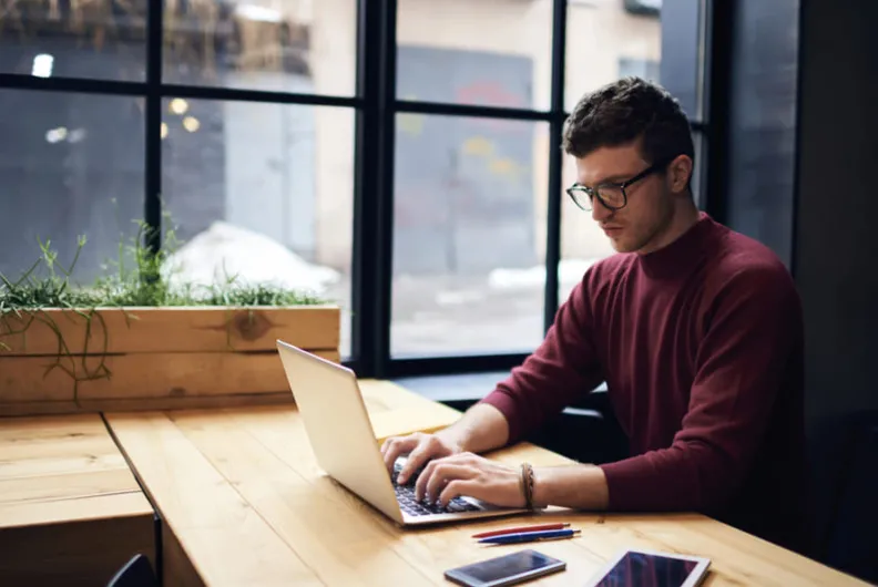 Man in glasses working on a laptop at a wooden desk by a window.