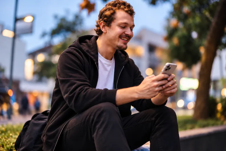 A smiling man sitting outdoors in an urban setting at dusk, using his smartphone, representing mobile-first digital communication and engagement.